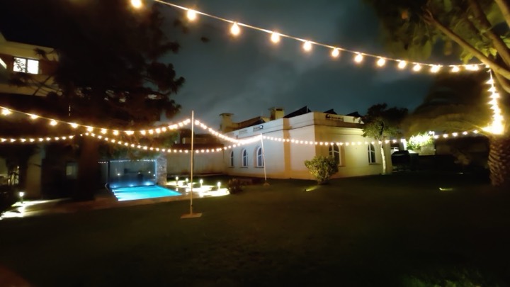 Guirnaldas cruzando jardín de casona con piscina iluminada, nocturna
